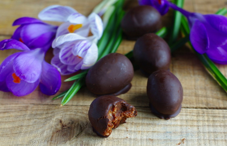 Small glossy dark chocolate Easter eggs, a bite taken from one, surrounded by purple and white flowers.