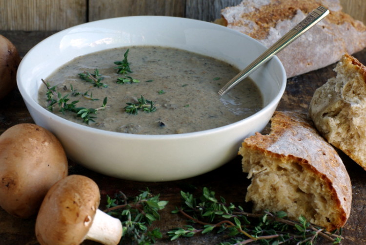 A bowl of earthy brown cream soup topped with green thyme leaves, and chunks of rustic bread on the side.